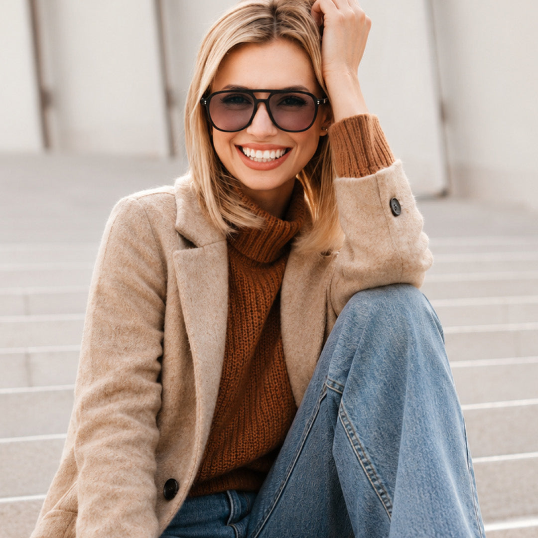 A smiling woman wearing black sunglasses, a beige coat, a rust-brown knit turtleneck, and blue jeans, sitting on outdoor concrete steps.